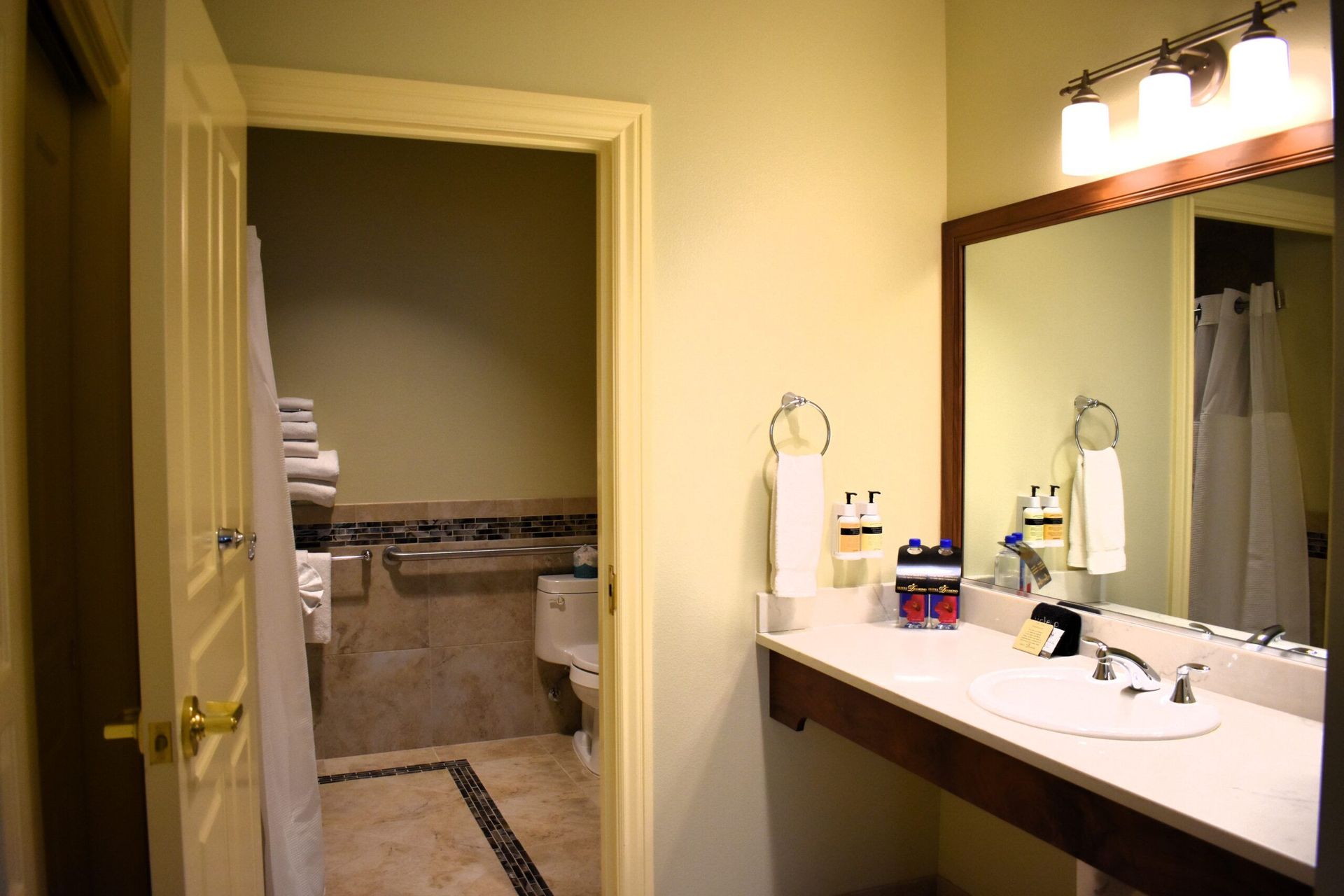 Bathroom with a vanity, mirror, and a doorway leading to a toilet and shower area. Beige walls and stone tile floor.