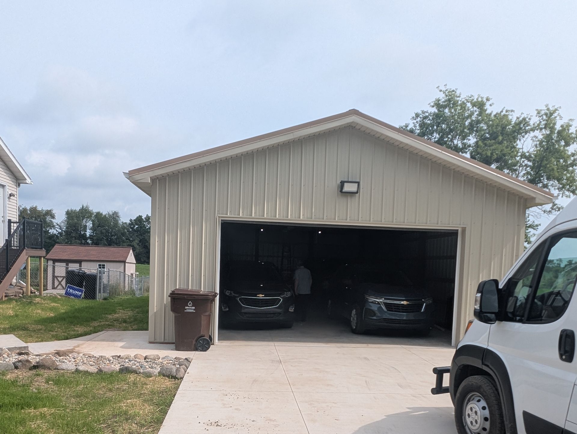 Detached beige garage with open doors, two cars parked inside, driveway in front, and a white van at right.