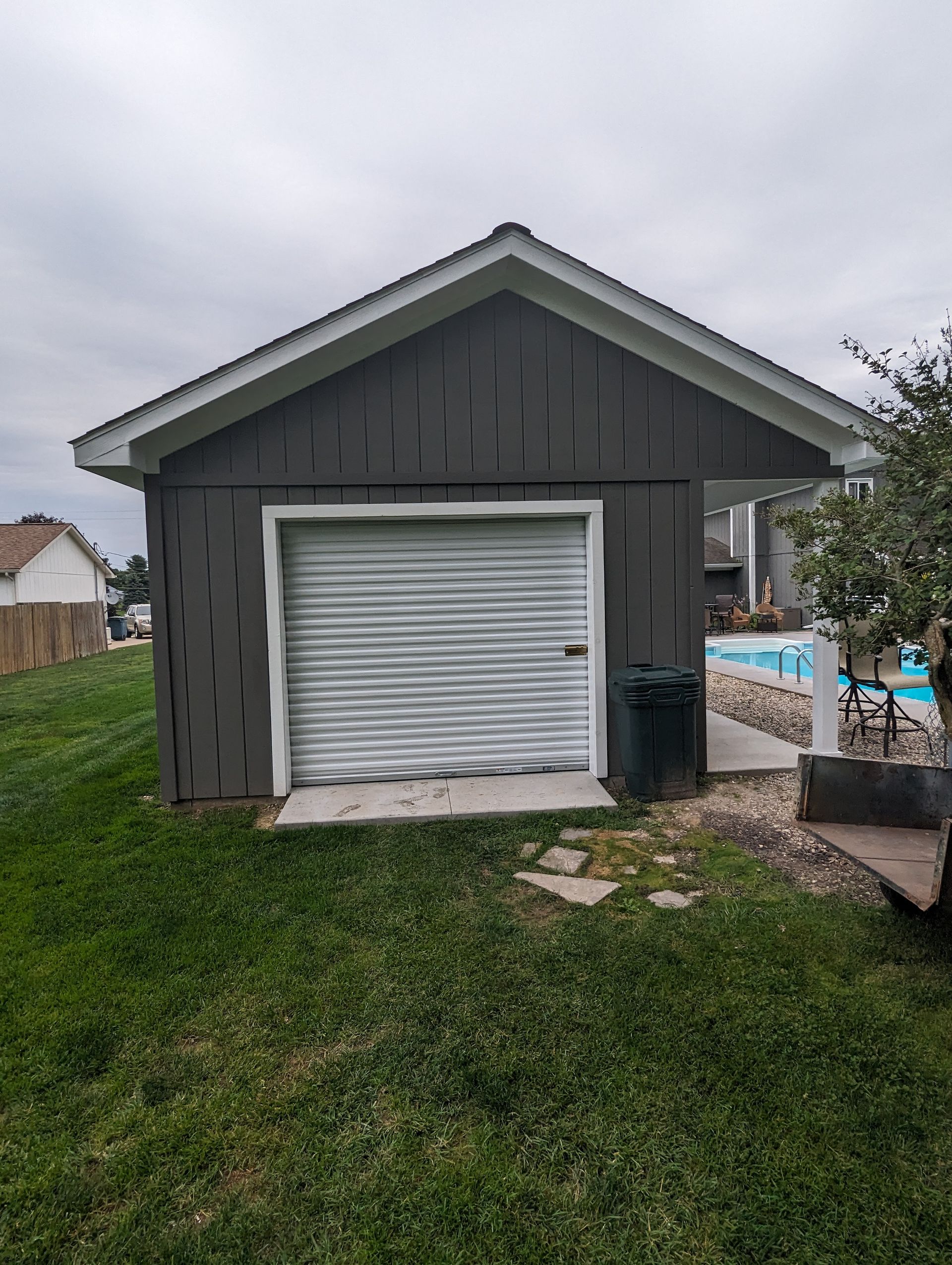 Gray shed with a white roll-up door beside a fenced backyard pool area under a cloudy sky