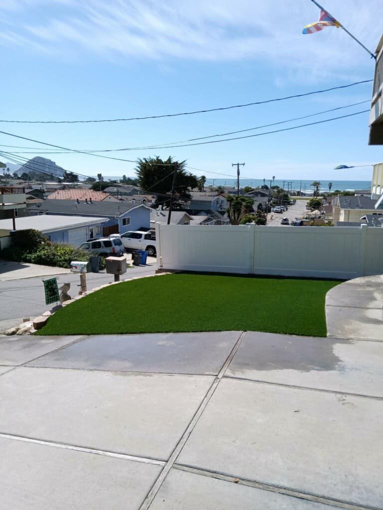 View from a driveway toward a white wall, green lawn, and distant ocean under a clear blue sky.