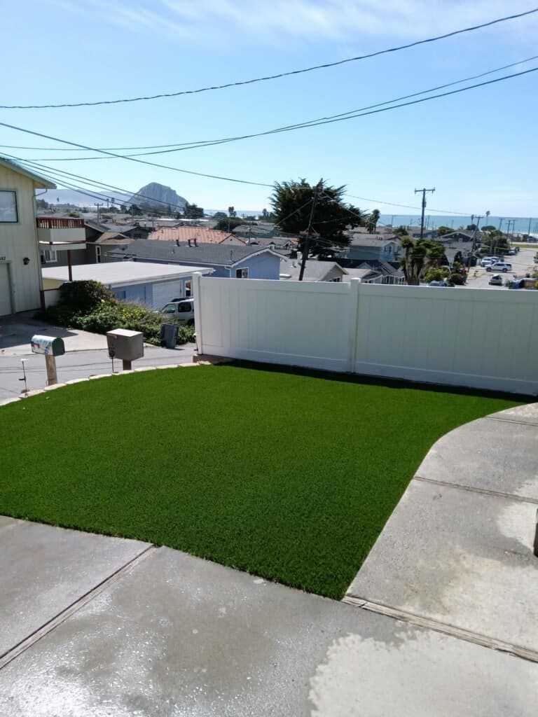 Backyard with bright green artificial grass, concrete patio, white wall, and ocean view beyond rooftops