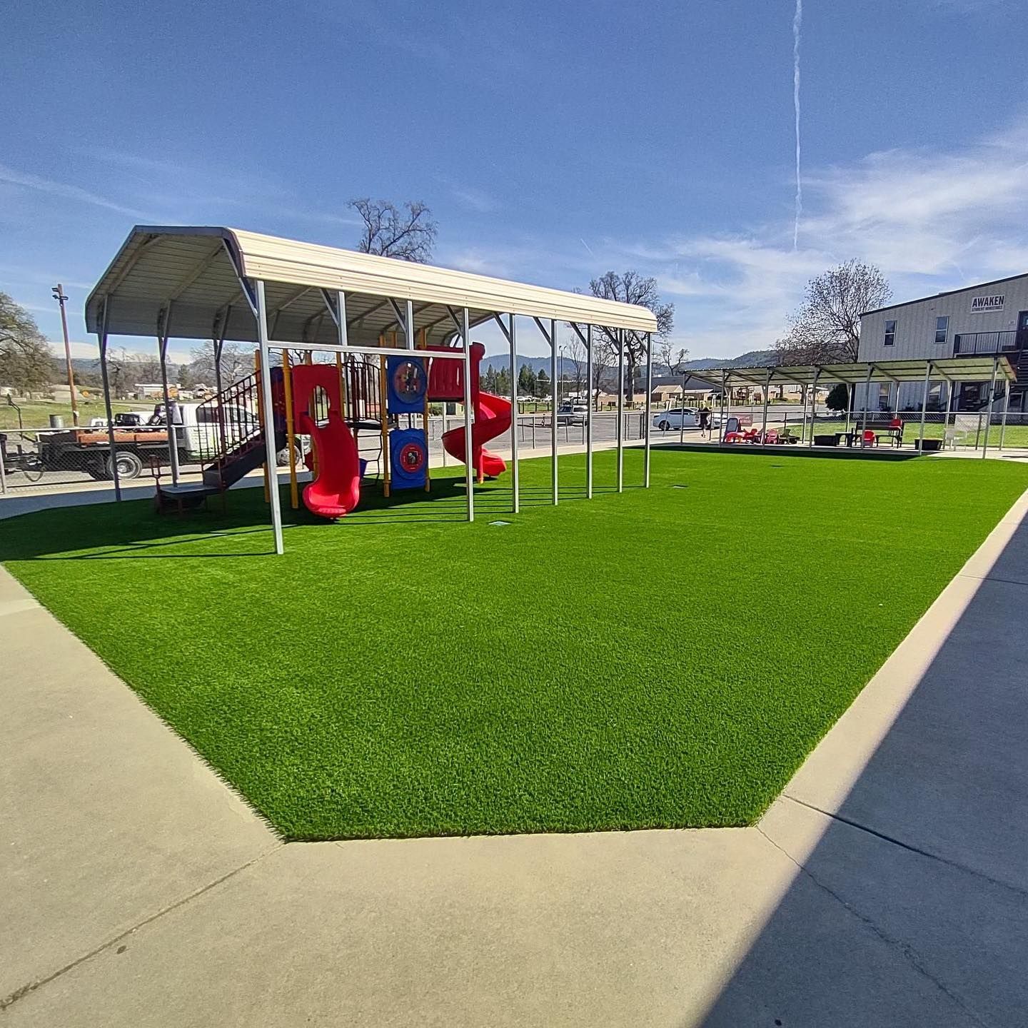 Covered playground with red and blue play structures on bright green turf in a sunny park