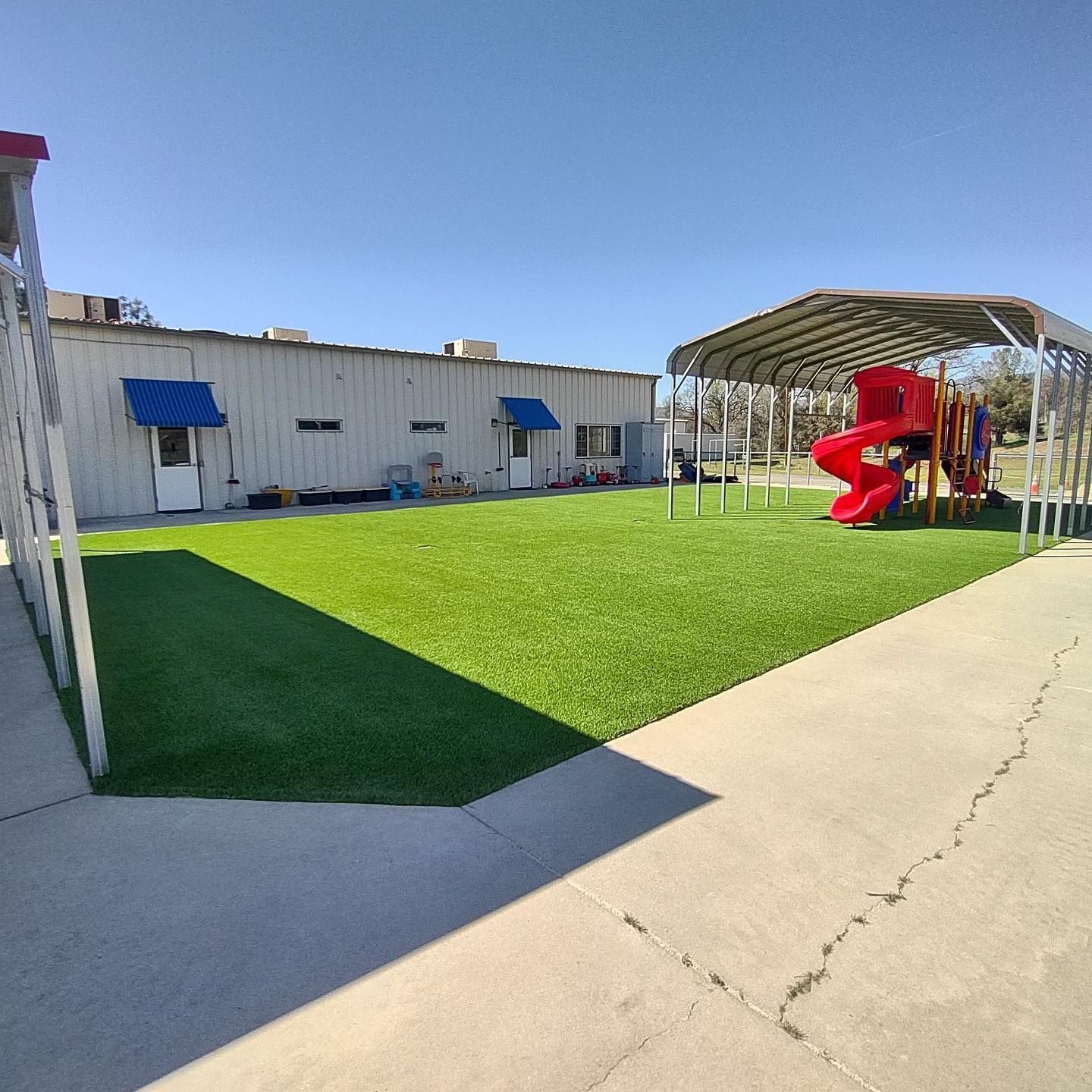 Sunny playground with green turf, red slide, and shaded canopy beside a concrete walkway