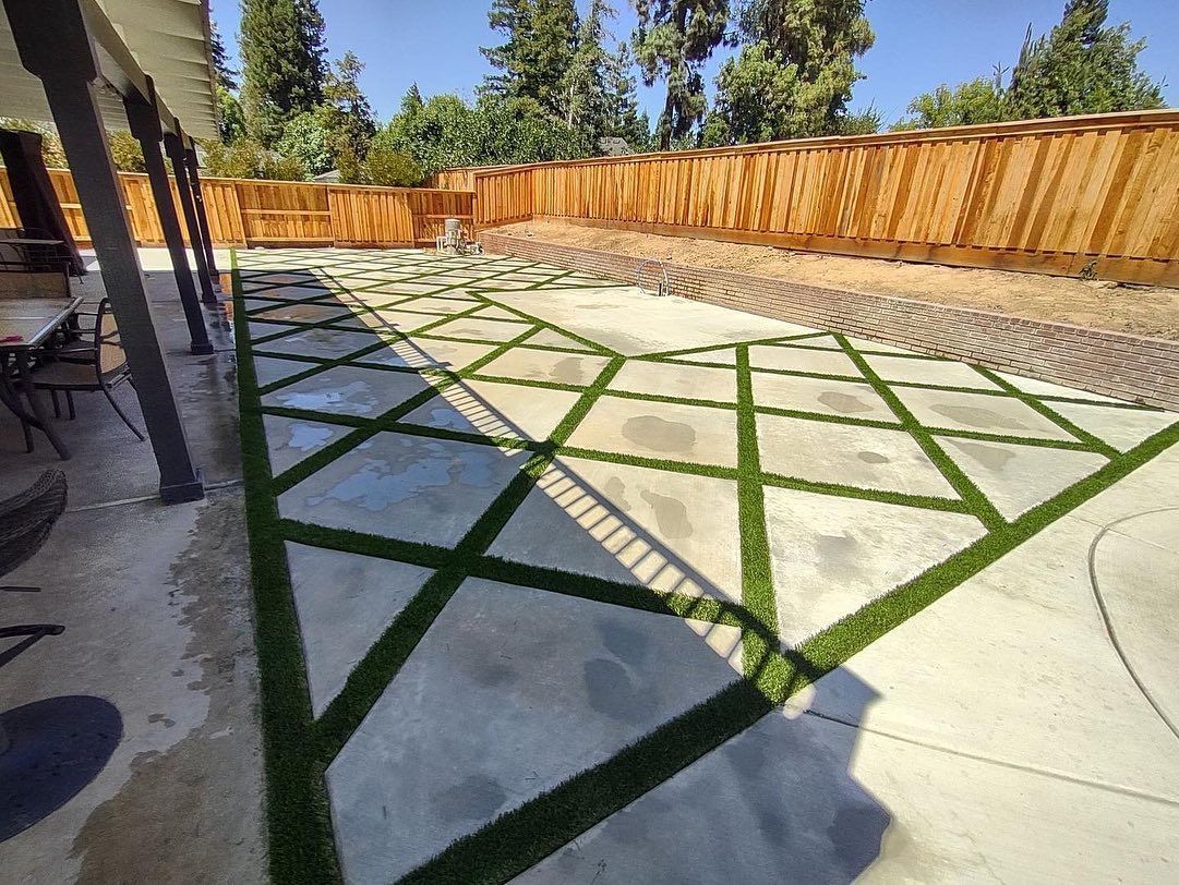 Backyard patio with geometric concrete pavers and green grass strips, bordered by a wooden fence.