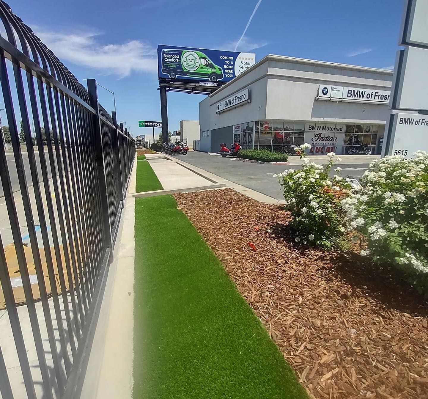 Sidewalk beside a fenced commercial building with a billboard, landscaped grass, and flower beds under a blue sky