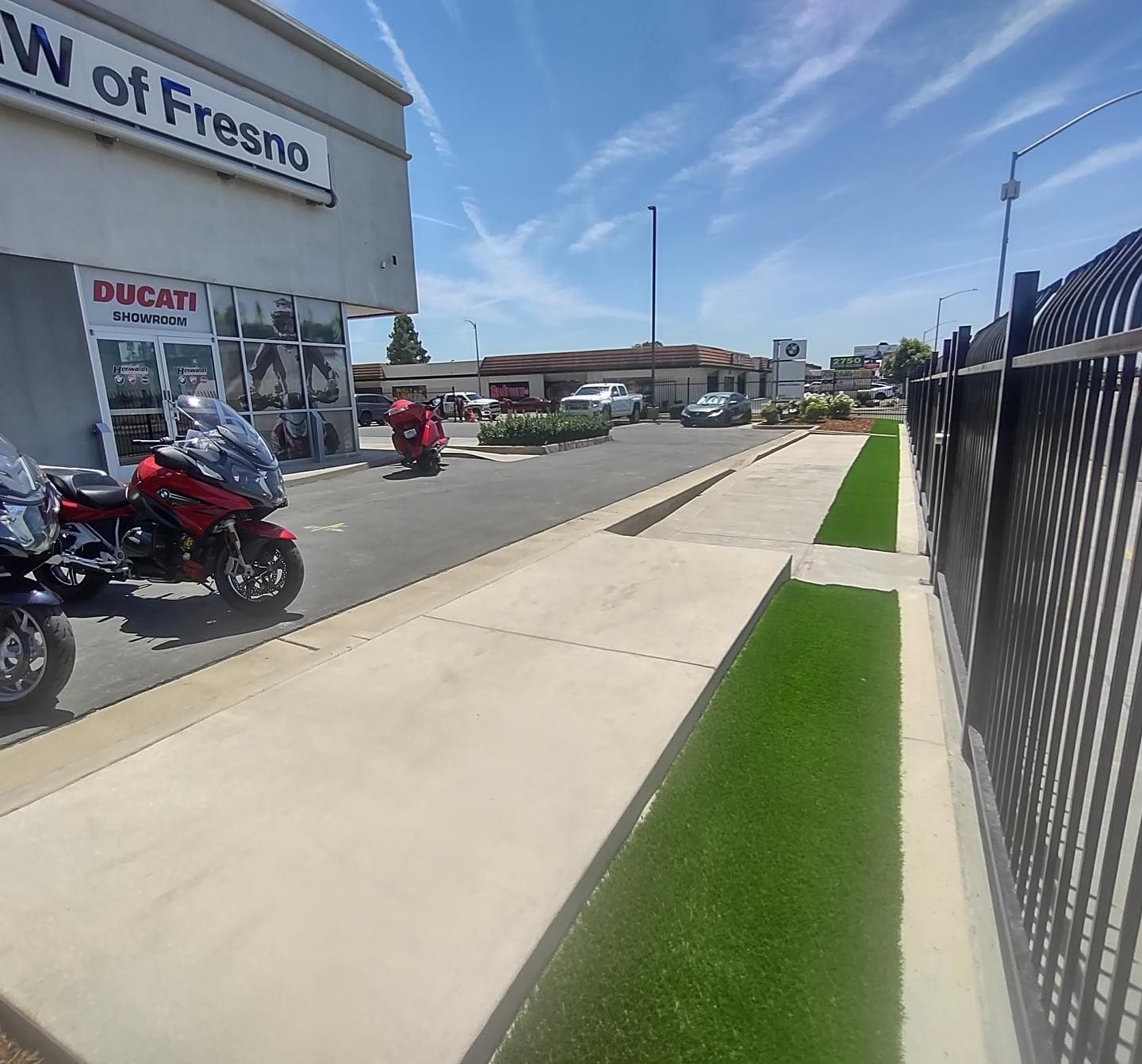 Sunny sidewalk beside a Ducati dealership, with parked motorcycles and a black fence along the street.