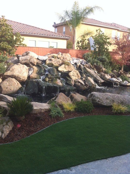 Landscaped backyard with stacked rocks, small waterfall, palm trees, and a green lawn near a house