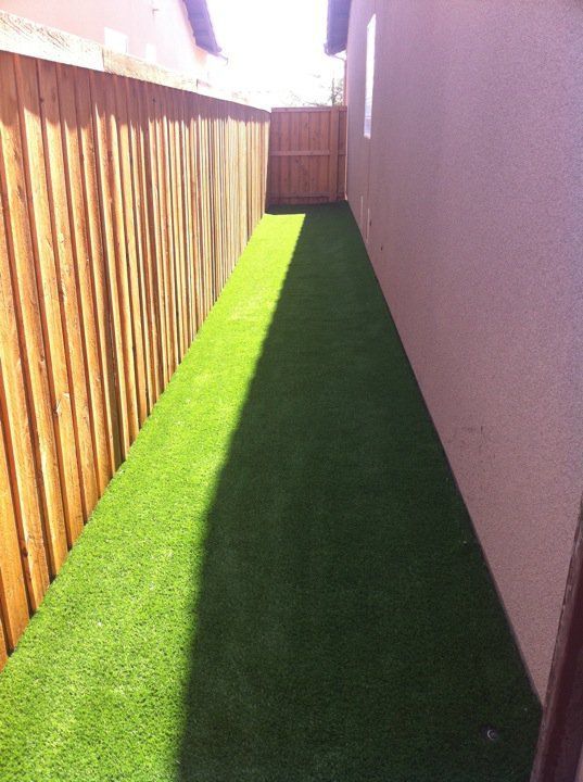 Narrow backyard walkway with green turf, wooden fence on one side and a house wall on the other.