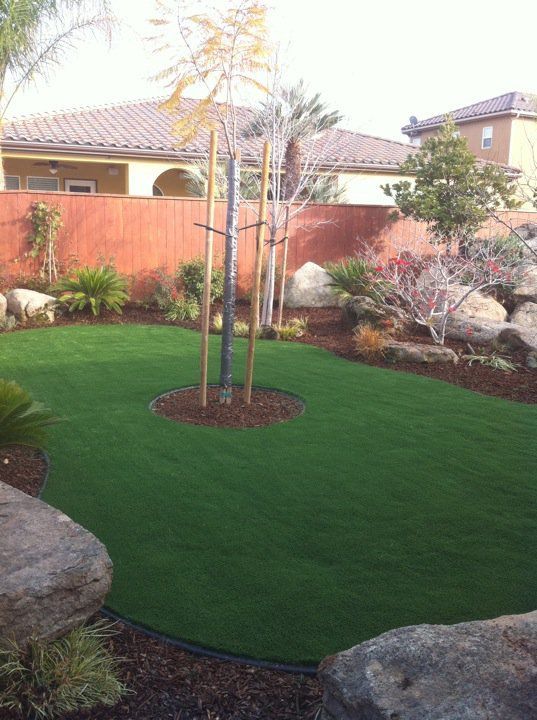 Backyard with a circular patch of green artificial turf around young trees, bordered by rocks and plants