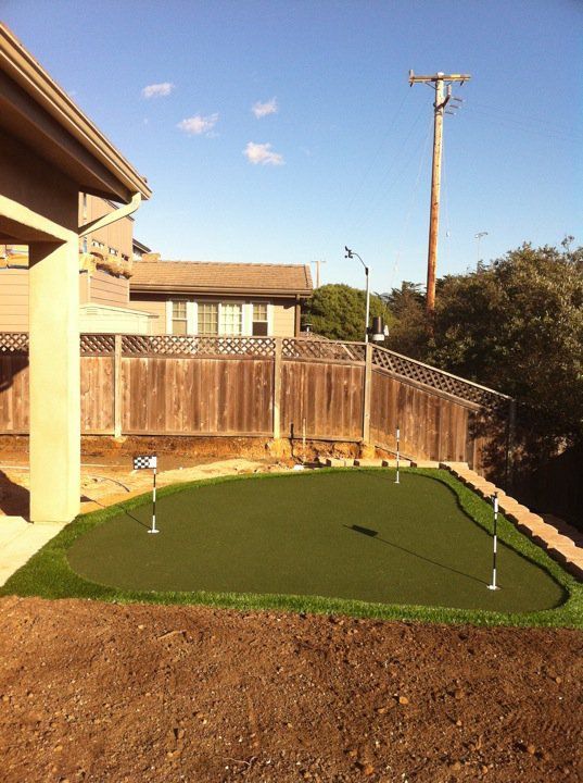 Backyard putting green with flagstick beside a wooden fence and house porch