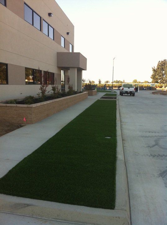 Sidewalk beside a beige building, with a narrow strip of bright green grass and a parking lot on the right.