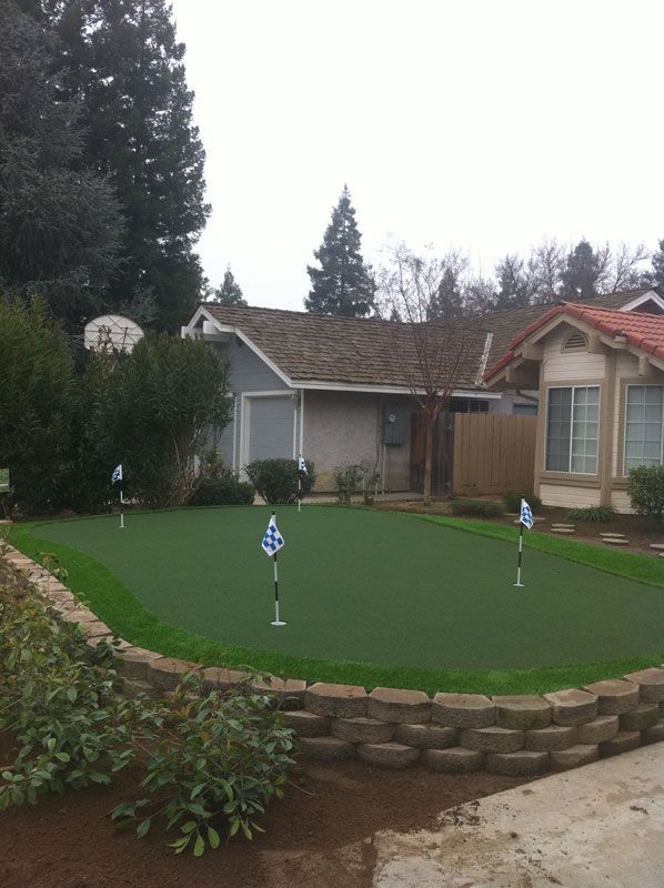 Backyard putting green beside a house, with two golf flags and a stone retaining wall.