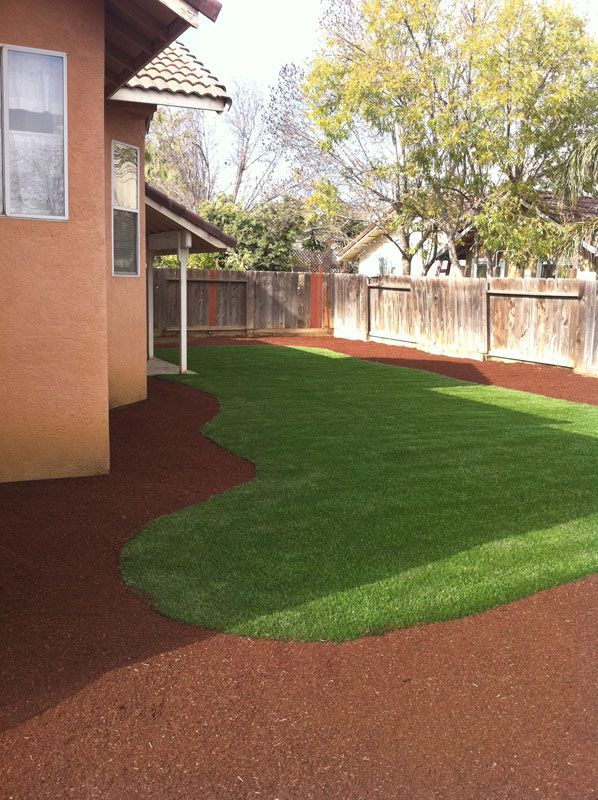 Backyard with green lawn, red gravel border, house wall, and wooden fence in daylight