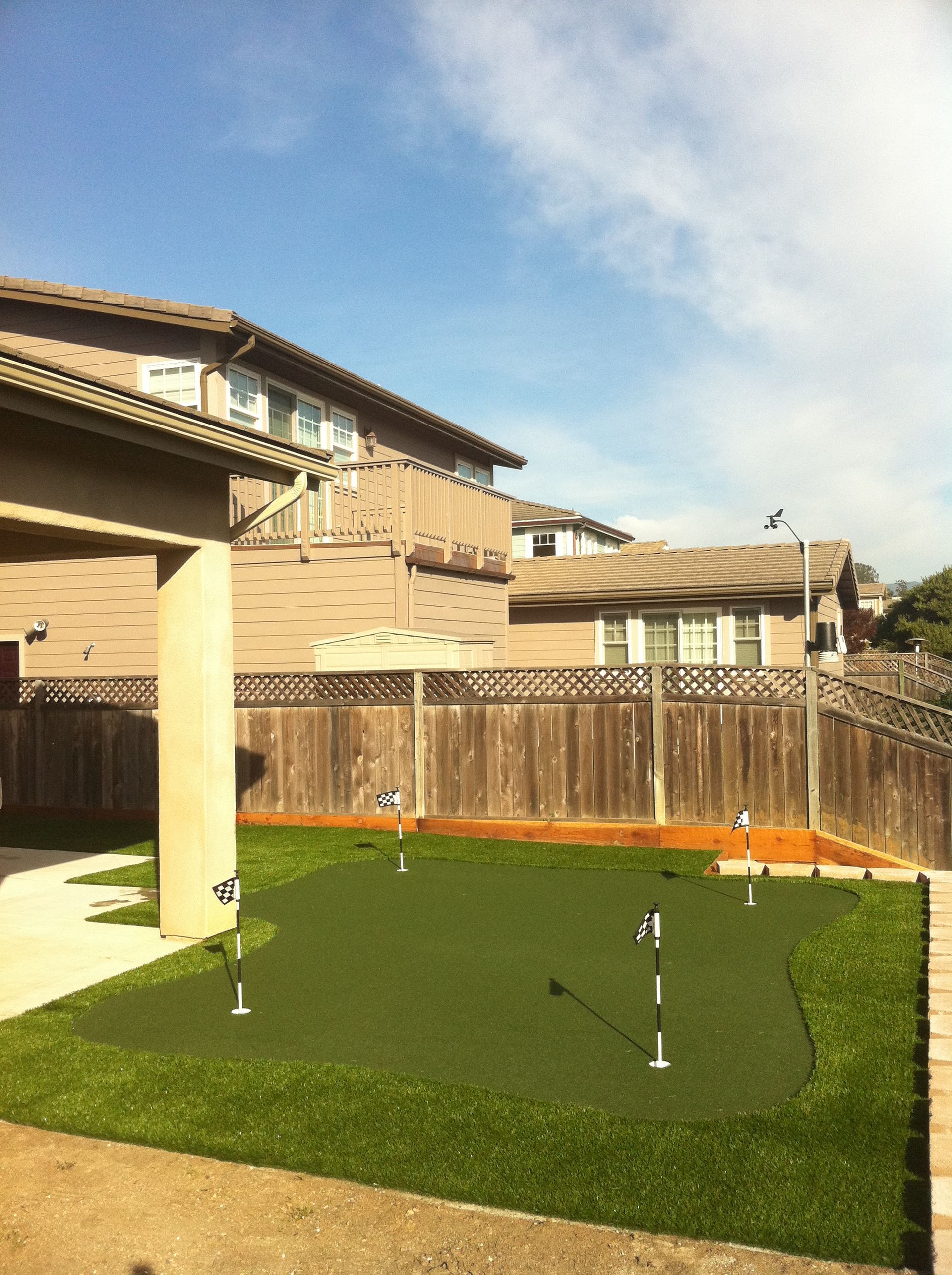 Backyard with green artificial turf, wooden fence, and two small soccer goal markers under a blue sky