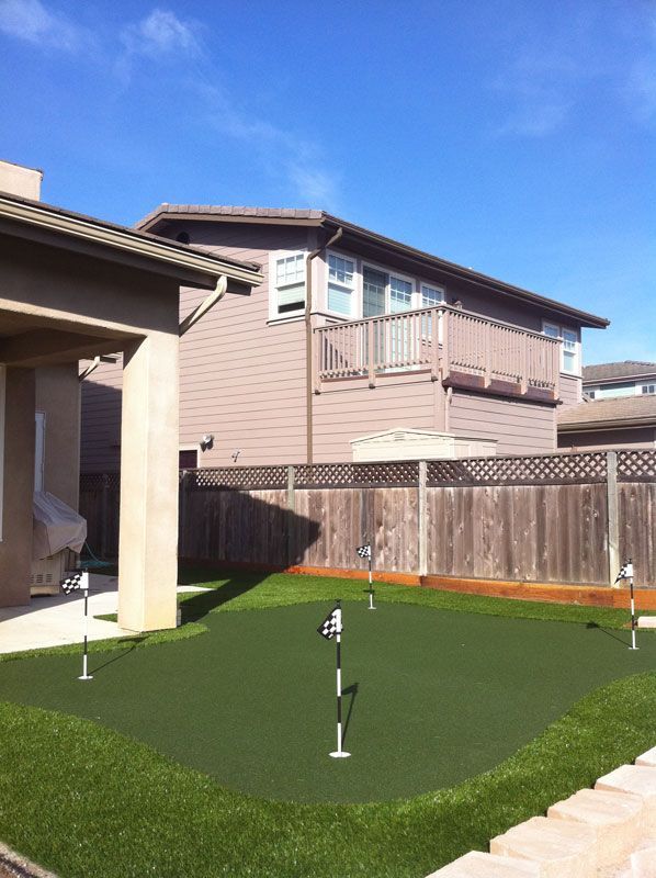 Backyard putting green with golf flags beside a house and wooden fence under a blue sky