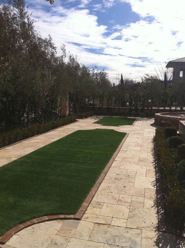 Stone patio with a curved green lawn strip, bordered by hedges and trees under a cloudy sky