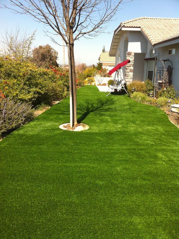 Freshly landscaped backyard with bright green artificial turf, a young tree, shrubs, and a patio chair by the house