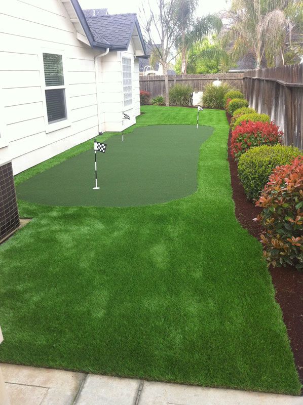 Backyard putting green with flagstick beside a house, bordered by shrubs and a walkway.