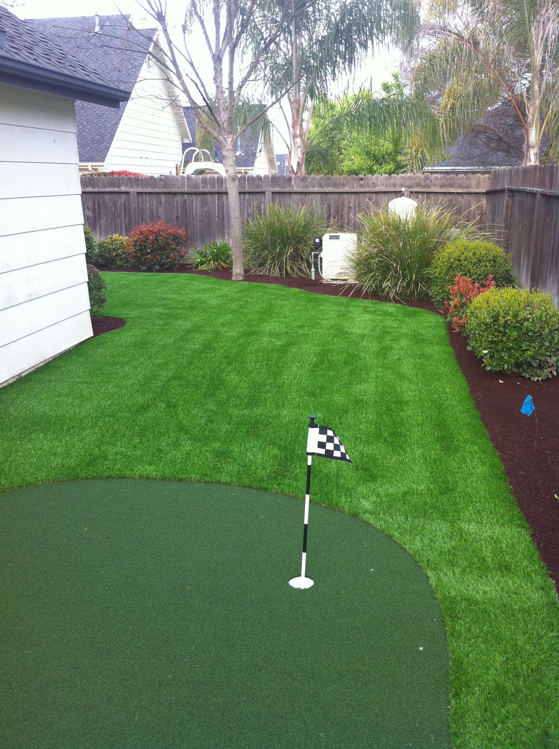 Backyard putting green with checkered flag, green turf, and trimmed shrubs beside a fence