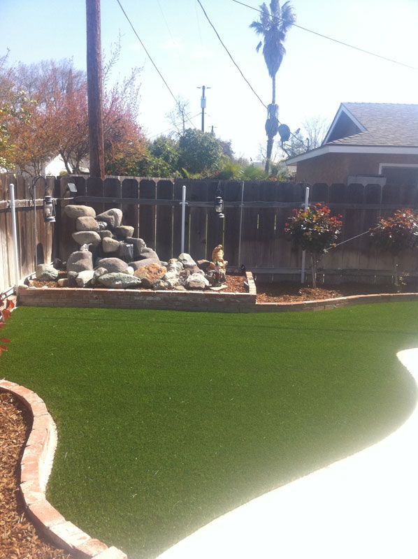 Backyard with artificial turf, rock waterfall fountain, and a fenced patio under a bright sky