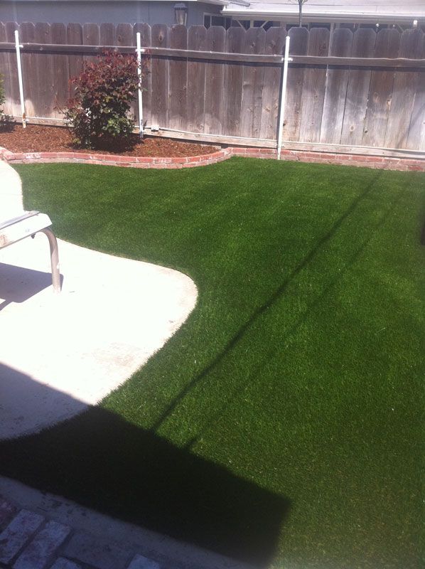 Backyard with bright green artificial turf, white patio area, fence, and a small bush in sunlight