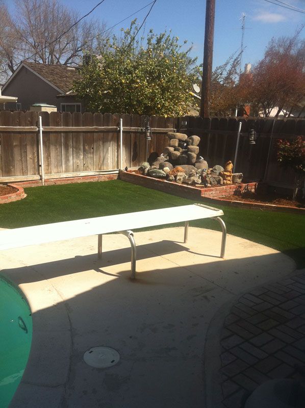 Backyard patio with a white table, green turf, wooden fence, and stacked rocks by the wall
