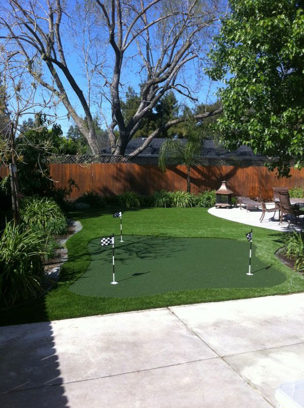 Backyard putting green with flags, bordered by patio, fence, and trees under a blue sky