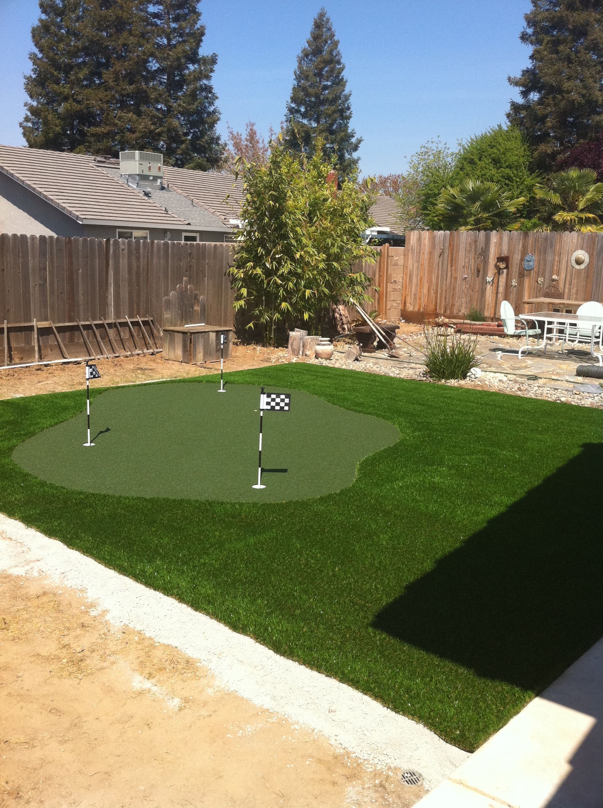 Backyard putting green with flagsticks on artificial turf, bordered by a wooden fence and trees