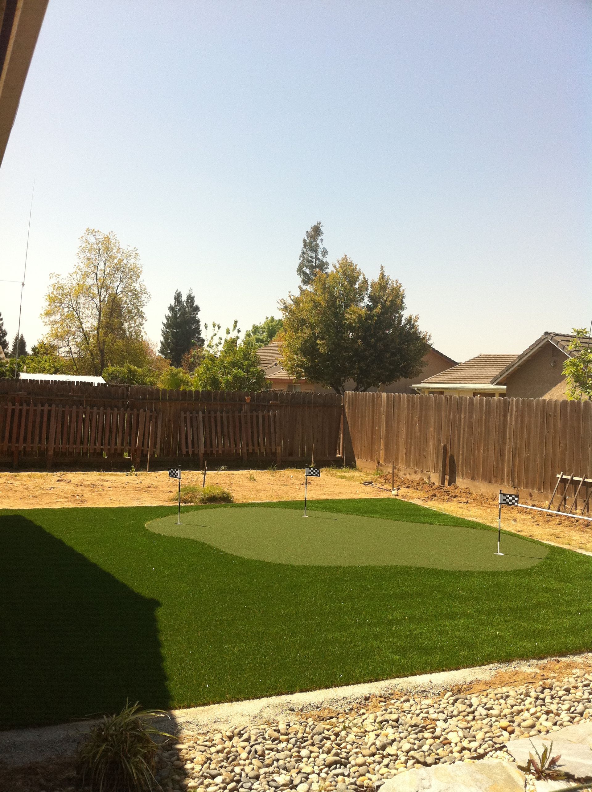 Backyard with green lawn, wooden fence, gravel border, and trees under a clear sky