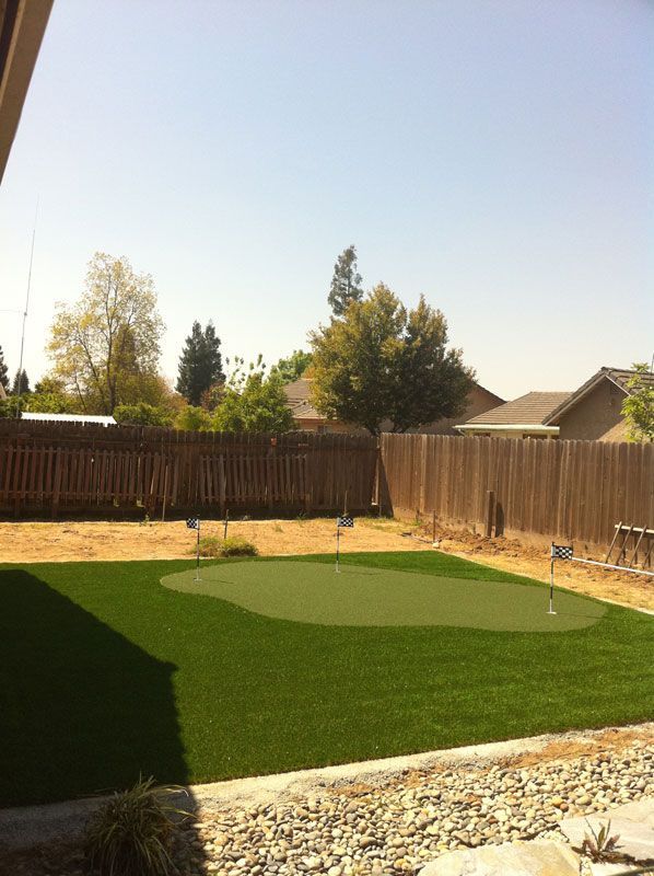 Backyard with artificial turf, wooden fence, gravel border, and trees under a clear sky