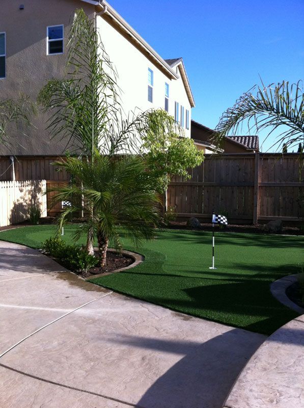 Backyard patio with artificial turf, palm tree, wooden fence, and a small putting green under a clear blue sky