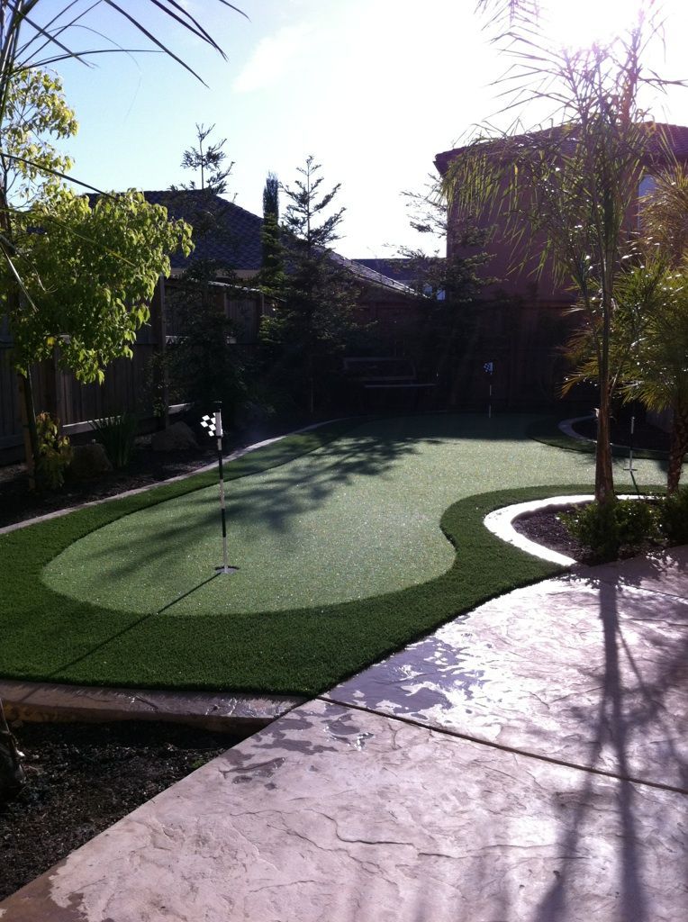 Backyard putting green with flag, surrounded by trees and a paved path in sunlight