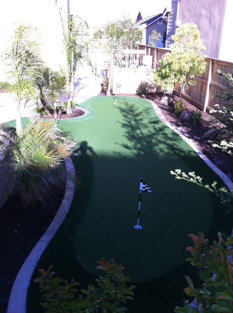 Backyard putting green with a golf hole, surrounded by plants and a fence