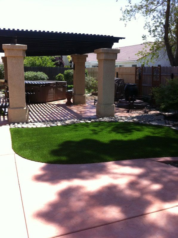 Backyard patio with pergola, stone columns, brick floor, green lawn, and a concrete walkway in sunlight