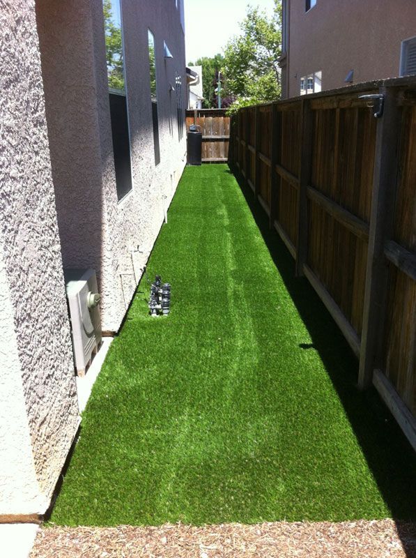 Narrow side yard with artificial turf between a stucco house wall and wooden fence, leading to a gate.