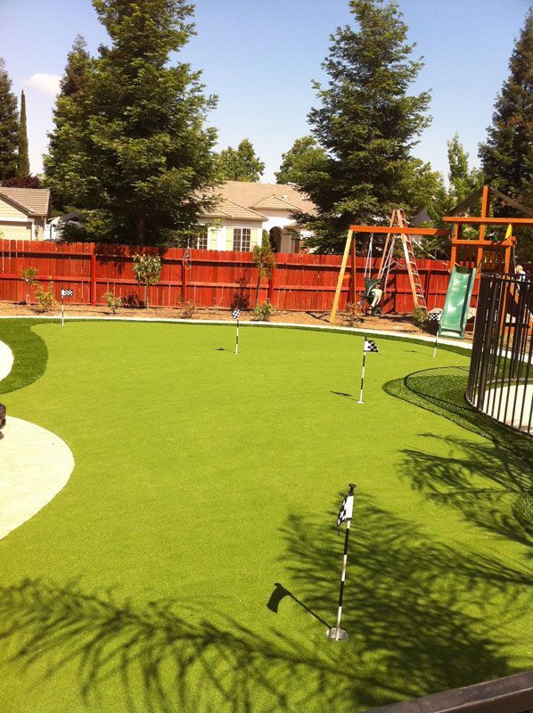 Backyard mini golf green with flagsticks, surrounded by red fence, trees, and a swing set