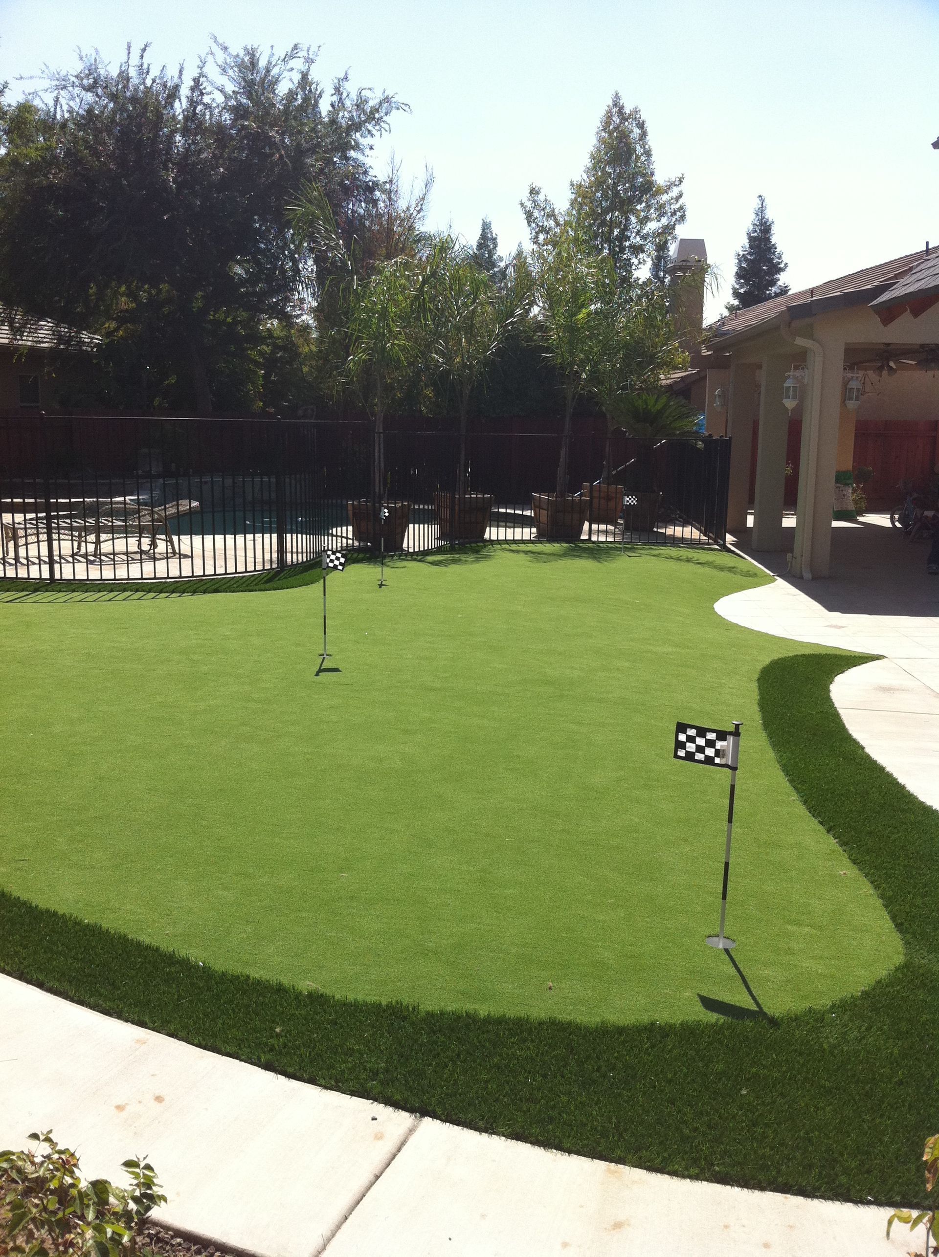 Backyard mini golf putting green with a flag, trees, and a patio in the sun