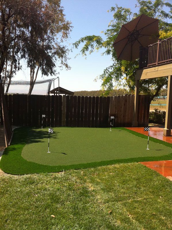 Backyard putting green beside a wooden fence, with a patio and umbrella in the corner.