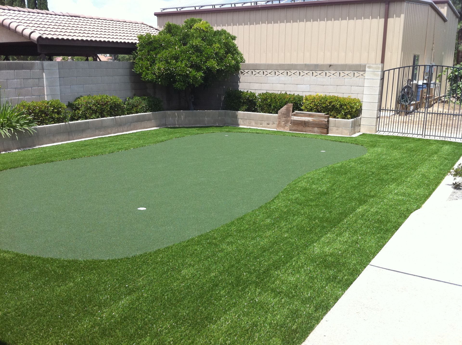 Backyard putting green with artificial turf, surrounded by grass, a wall, and a paved patio