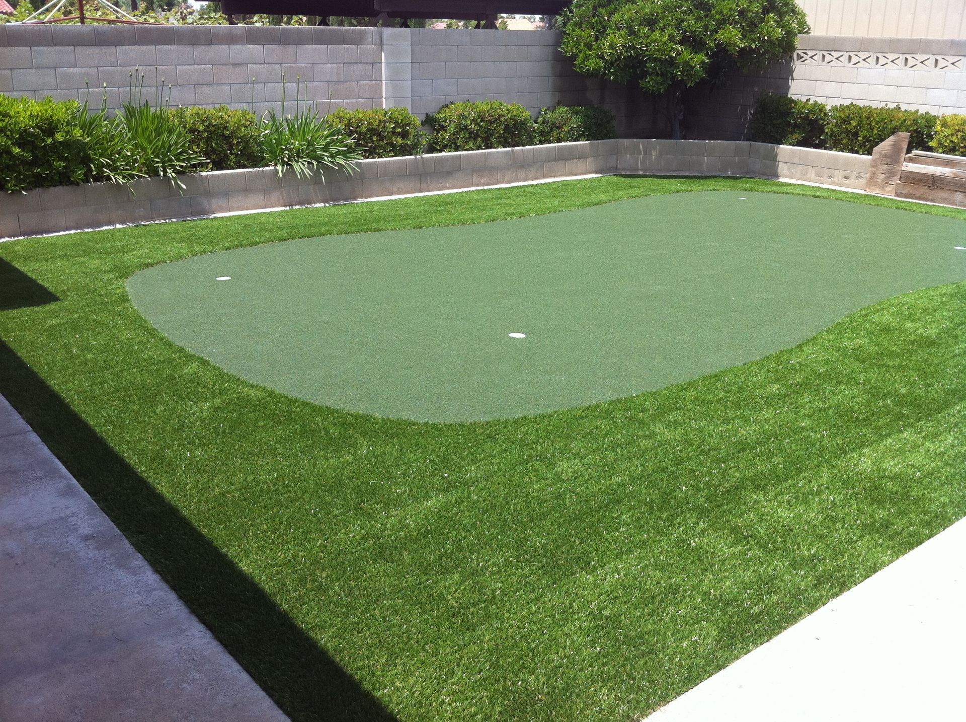 Backyard putting green with artificial turf, bordered by grass and a gray stone wall.