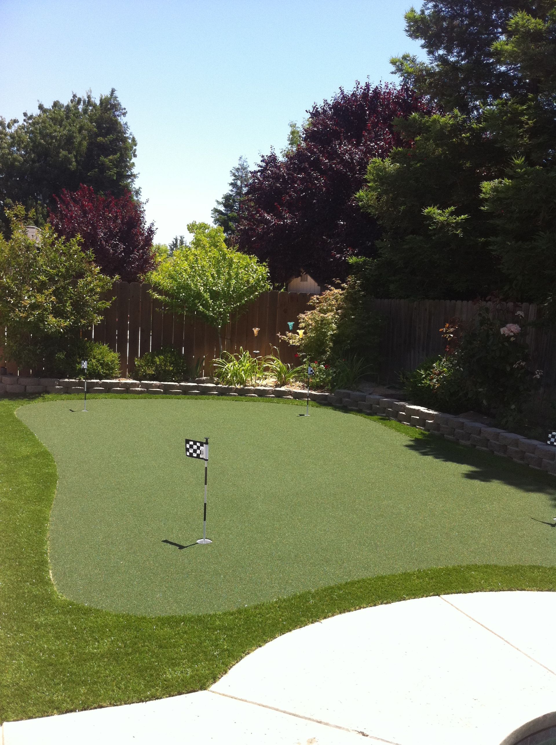 Backyard putting green with a golf flag, bordered by trees and a white patio edge