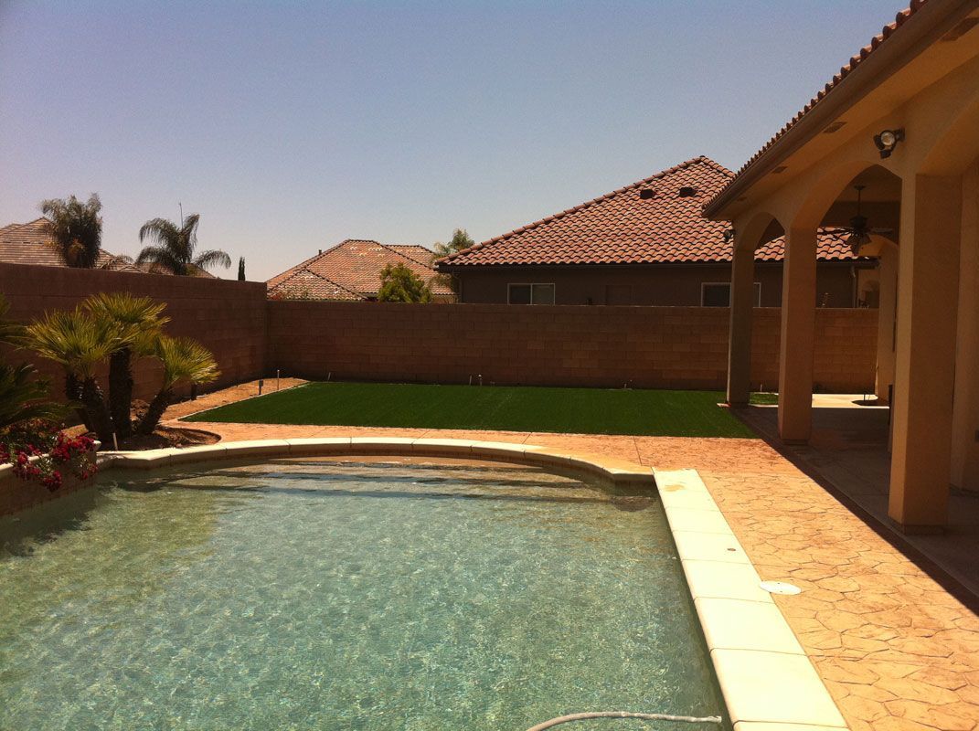Backyard swimming pool beside a patio and house, with a green lawn and tiled roof.