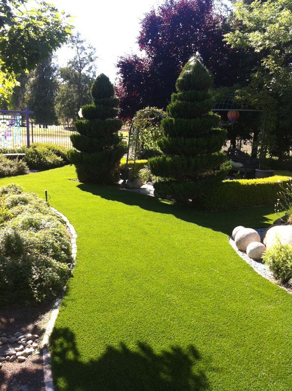 Sunlit landscaped garden with manicured topiary trees, bright green lawn, and decorative white rocks.