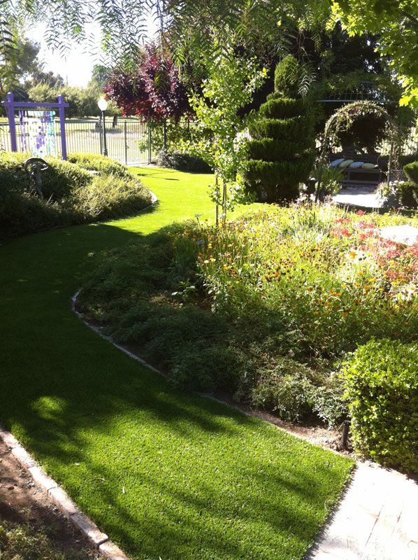 Sunny garden with green lawn, flower beds, shrubs, and a pathway near a fence