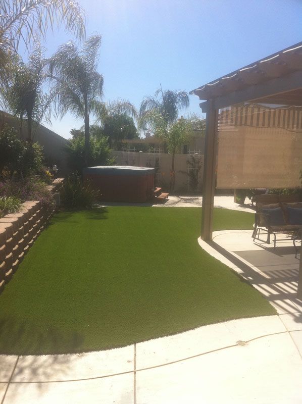 Sunny backyard patio with artificial grass, palm trees, and a covered seating area beside a hot tub.
