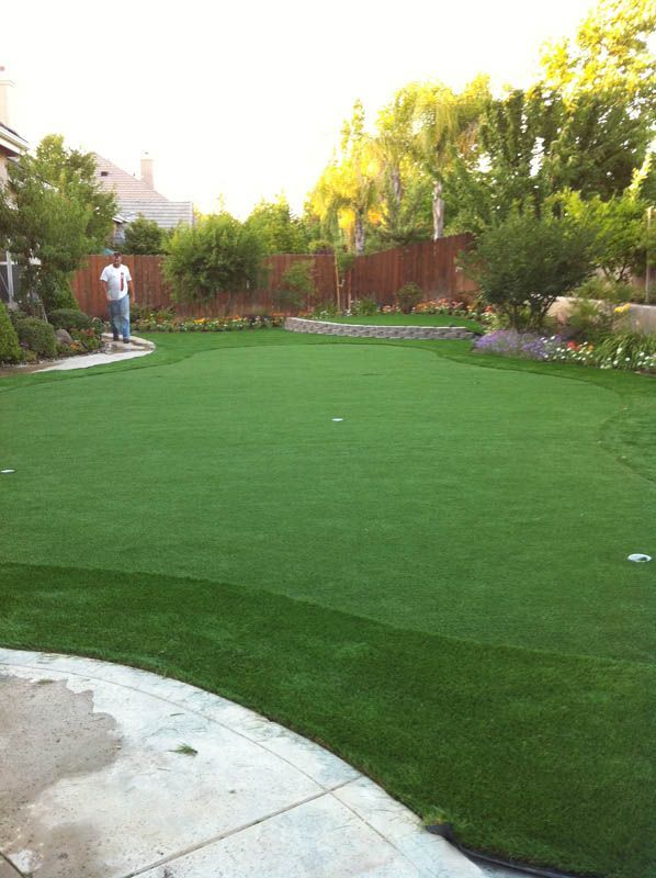 Green backyard lawn with curved stone patio, flower beds, trees, and a person standing near the fence