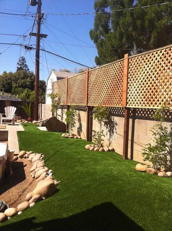 Landscaped backyard with artificial turf, stone edging, lattice fence, and tall trees under a blue sky