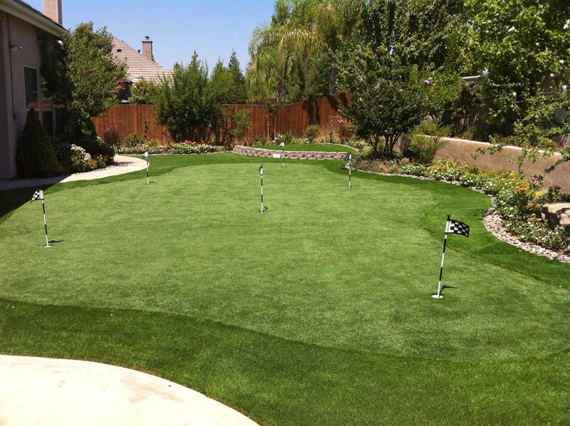 Backyard putting green with golf flags, trees, and flower beds in bright sunlight