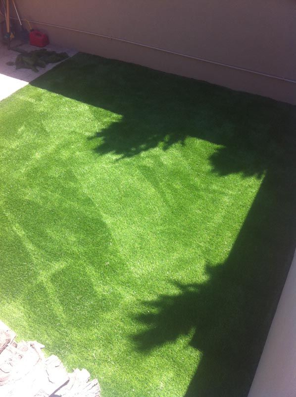 Sunlit green artificial grass mat in a corner, with shadows from nearby plants and walls.