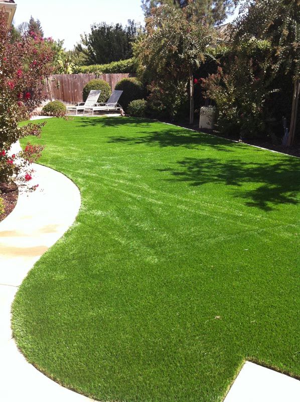 Curved, lush green lawn in a landscaped backyard with patio chairs and trees under bright sunlight
